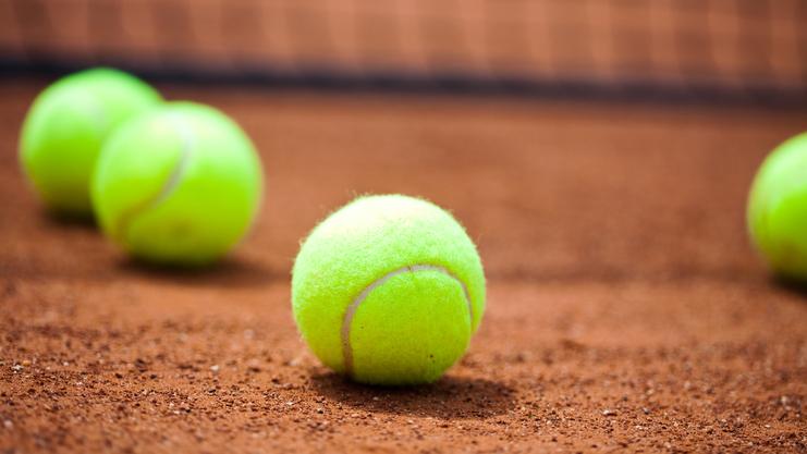 Vier grüne Tennisbälle liegen auf einem roten Sandplatz, mit einem Netz im Hintergrund.