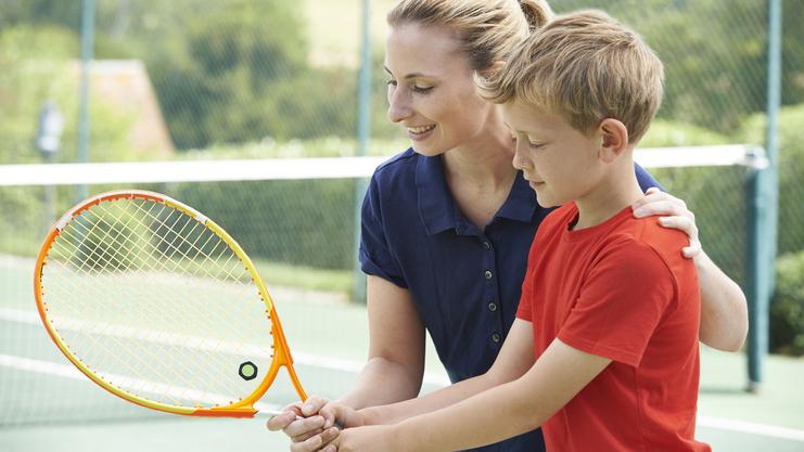 Eine Frau hilft einem Jungen beim Halten eines Tennisschlägers auf einem Tennisplatz.