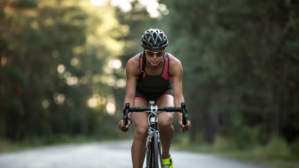 Sportlerin auf Rennrad in sportlicher Bekleidung fährt auf asphaltierter Straße durch bewaldetes Gebiet.