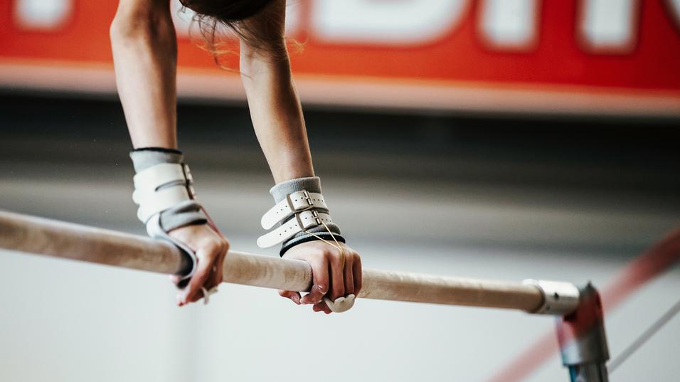 Nahaufnahme zweier Hände, die eine schmale Turnstange mit Griffpolstern halten, im Hintergrund eine Turnhalle.