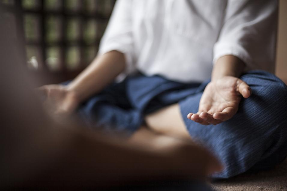 Nahaufnahme einer sitzenden Person in meditativer Haltung mit ausgestreckten Händen auf einem Teppichboden.
